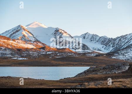 Un paysage panoramique à couper le souffle avec des montagnes enneigées et un lac calme reflétant le ciel clair et la lumière de l'heure dorée Banque D'Images
