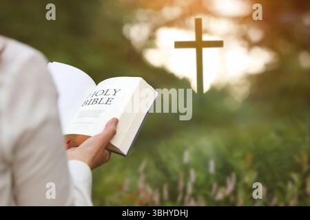 Femme chrétienne tenant la Bible et lisant la parole de Dieu dans la forêt avec la croix d'église brillamment éclairée, la foi et la croyance fond Banque D'Images