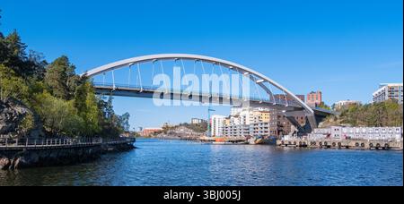 Stockholm, Suède - 5 mai 2025 : un pont en arc moderne enjambant une rivière calme, entouré d'arbres et de bâtiments urbains. Le ciel est clair et bleu, Banque D'Images