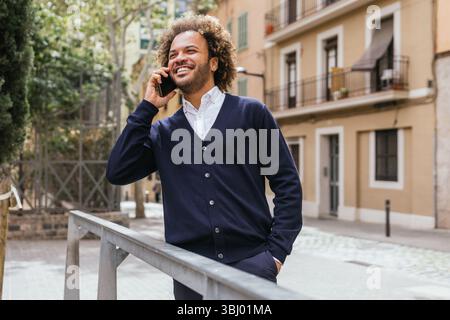 Un homme souriant prend une pause du travail dans un espace de coworking à Barcelone, en utilisant son smartphone à l'extérieur. La scène capture un moment de détente au milieu Banque D'Images