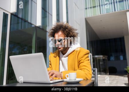 Un homme d'affaires aux cheveux afro, portant des lunettes de soleil et un manteau jaune, aime travailler sur son ordinateur portable tout en étant assis à l'extérieur à une table avec une tasse à café, Banque D'Images