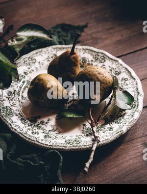 Trois poires mûres reposent sur une assiette à motifs complexes, placée sur une table en bois rustique. L'éclairage naturel améliore les tons chauds et terreux, créant un Banque D'Images