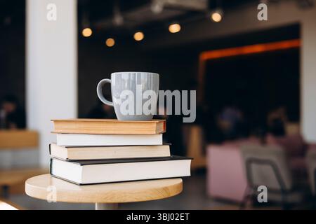 Une scène de café sereine avec une pile de livres et une tasse grise sur une table en bois, sur fond d'un intérieur légèrement flou et lumineux chaud Banque D'Images