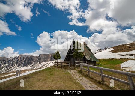 Une cabane rustique En A avec un toit vert se dresse sur une colline herbeuse à Passo Giau, entourée de plaques de neige et de spectaculaires sommets Dolomites sous un c dynamique Banque D'Images