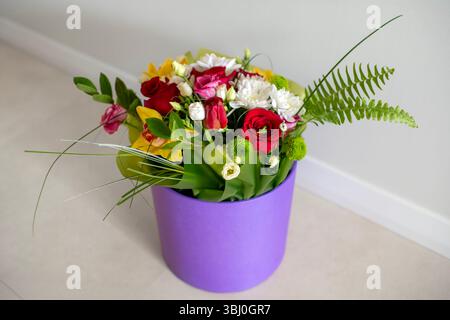 Un bouquet lumineux de fleurs mélangées comprenant des roses, des tulipes, des chrysanthèmes et de la verdure dans un coffret cadeau rond violet, vue de dessus, sur un fond clair. Banque D'Images