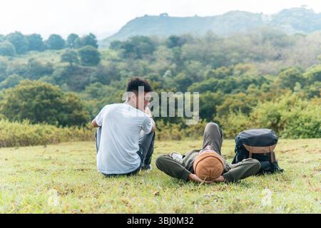 Deux hommes indonésiens se détendent sur une colline herbeuse, riant et dégustant une tasse de café avec une forêt pittoresque et une montagne en toile de fond. Banque D'Images