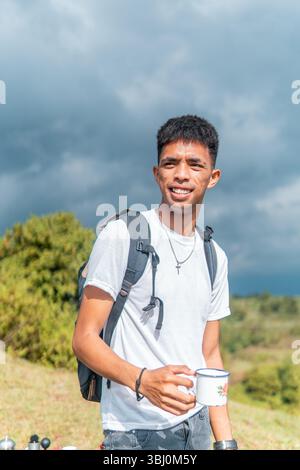 Un homme indonésien avec un sac à dos et un bonnet déguste une tasse de café tout en regardant au loin. Banque D'Images