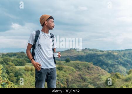 Un homme indonésien avec un sac à dos et un bonnet déguste une tasse de café tout en regardant au loin. Banque D'Images