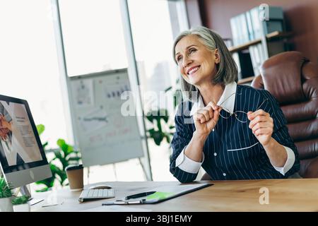 Élégante femme d'affaires mature dans un bureau moderne tenant des lunettes et souriant avec confiance tout en s'engageant dans des tâches professionnelles Banque D'Images