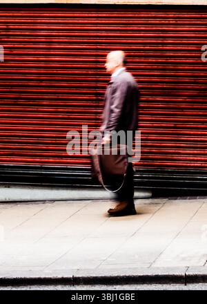Image floue d'un homme marchant sur un trottoir, portant une mallette, devant un volet rouge. Banque D'Images