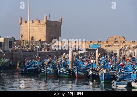 Essaouira, Maroc - 21 février 2025 : des bateaux de pêche traditionnels sont ancrés dans le port d'Essaouira avec la tour de la fortification de Mogador à Mor Banque D'Images