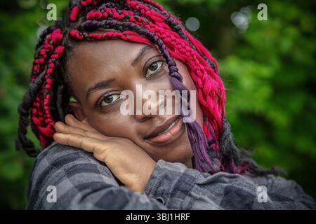 Charmante femme afro-américaine avec coiffure tressée rose reposant la tête sur les mains dans le parc vert. Portrait naturel du style de vie en extérieur avec un sourire authentique Banque D'Images