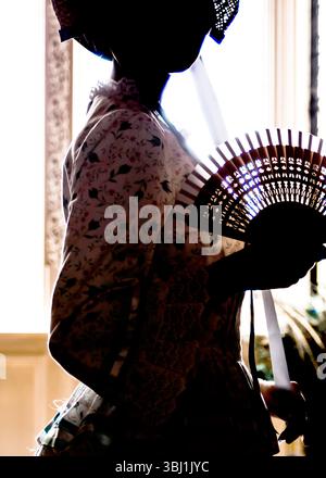 Une personne en silhouette, portant un capot et tenant un ventilateur, se tient près d'une fenêtre. Banque D'Images