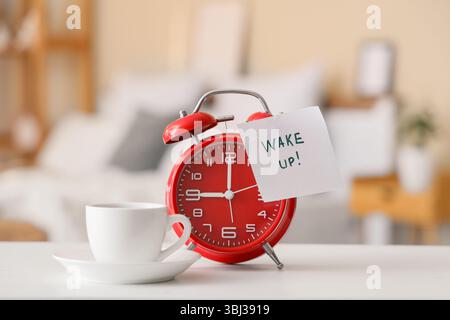 Réveil, tasse de café et note collante avec texte RÉVEILLER sur la table de chevet dans la chambre, gros plan Banque D'Images