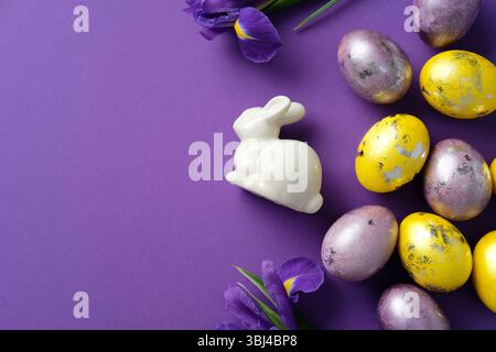 Composition de pose à plat élégante de Pâques. Lapin en céramique, œufs violets et jaunes, et iris frais sur fond violet Banque D'Images