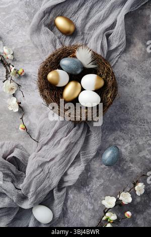 Composition de Pâques avec des œufs dorés, blancs et bleus mouchetés dans un nid, entouré de branches en fleurs et de tissu gris Banque D'Images