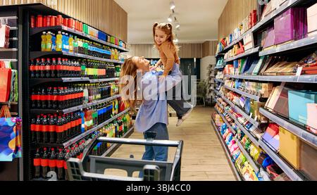 Maman joyeuse soulevant sa fille avec des rires dans l'allée du supermarché. Joyeux moment en famille lors de l'épicerie. Amour, lien, parentalité et joie Banque D'Images