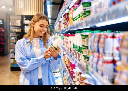 Femme souriante lit l'étiquette sur une bouteille de produit laitier tout en faisant ses courses dans une épicerie. Concept de consommateur conscient, mode de vie sain, nutrition, p Banque D'Images