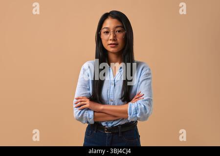 Une jeune femme joyeuse et énergique portant des lunettes élégantes pose avec confiance, les bras croisés, mettant en valeur un sourire chaleureux et invitant contre un b neutre Banque D'Images
