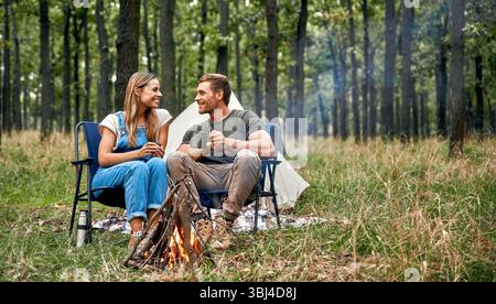Un couple profite d'un moment agréable et joyeux autour du feu de camp chaud dans une forêt magnifiquement sereine, créant des souvenirs inoubliables et durables im Banque D'Images