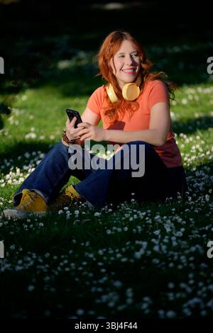Femme rousse à l'aide d'un smartphone assis dans l'herbe portant des écouteurs souriant dans un parc Banque D'Images