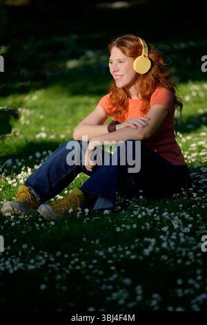 Étudiant Redhead appréciant la musique avec des écouteurs jaunes assis dans un champ d'herbe pendant une journée ensoleillée Banque D'Images