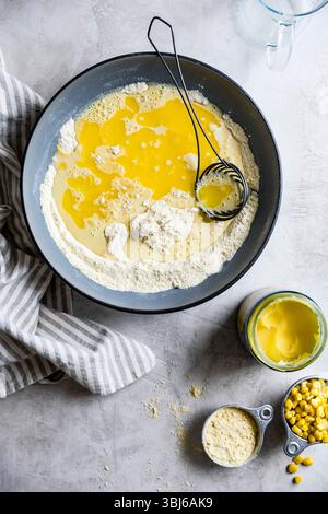 Une vue aérienne d'un bol à mélanger avec de la farine, des œufs et d'autres ingrédients en cours de préparation pour la cuisson, à côté du maïs et du beurre sur un comptoir de cuisine. Banque D'Images