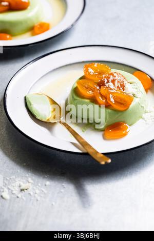 Une assiette de panna cotta verte garnie de tranches de kumquats et d'une pincée de copeaux de chocolat blanc, accompagnée d'une cuillère en or. Banque D'Images