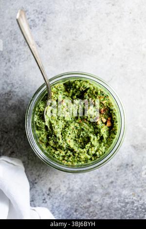 Un pot de pesto vert avec une cuillère sur une surface texturée. Banque D'Images