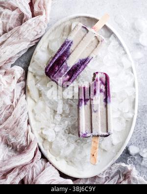 Deux popsicles violettes et crème sur un lit de glace dans un plat blanc, avec un chiffon rose à proximité. Banque D'Images