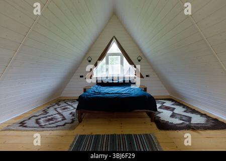 Cette image présente une chambre minimaliste à cadre En A avec un design en bois, avec un lit moelleux, des tapis élégants et une grande fenêtre pour laisser entrer la lumière naturelle Banque D'Images