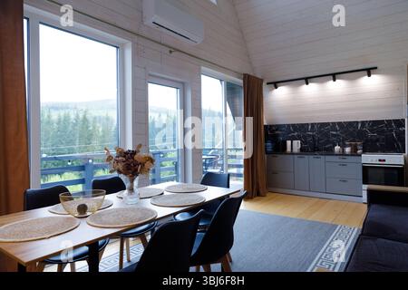 Cette image présente une cuisine moderne et un espace salle à manger avec un mobilier élégant, de grandes fenêtres révélant une vue panoramique sur la montagne, et un atmo serein Banque D'Images