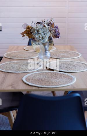 Une charmante table de salle à manger avec des tapis de table tissés, avec un vase en verre délicat rempli d'un assortiment de fleurs séchées, créant une atmosphère sereine et sereine Banque D'Images