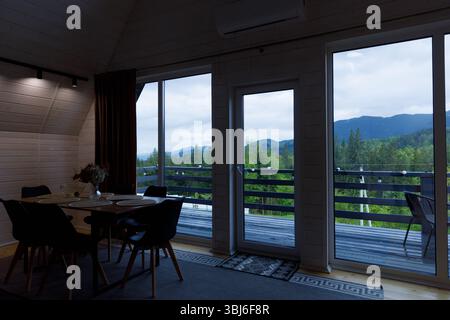 Un intérieur de cabine en bois invitant mettant en vedette une salle à manger avec des chaises élégantes, de grandes fenêtres panoramiques révélant des montagnes verdoyantes et un cadre tranquille Banque D'Images