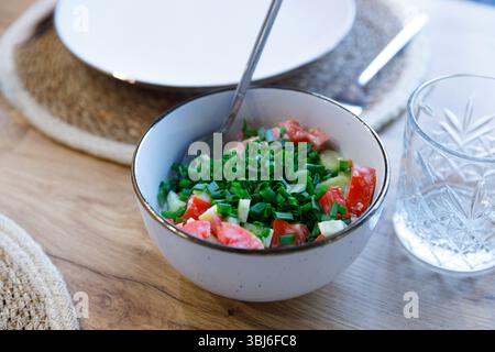 Cette image présente une salade vibrante avec des tomates coupées en dés et des oignons verts finement hachés dans un bol élégant à côté d'un verre d'eau, créant un Banque D'Images