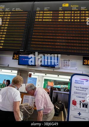 Enregistrement des passagers de la compagnie aérienne à l'aéroport couple mature avec leurs bagages à l'enregistrement au terminal de l'aéroport espagnol, avec des panneaux d'information de vol derrière, la manutention des bagages pour l'enregistrement sur leur vol British Airways de retour d'Espagne UE Banque D'Images