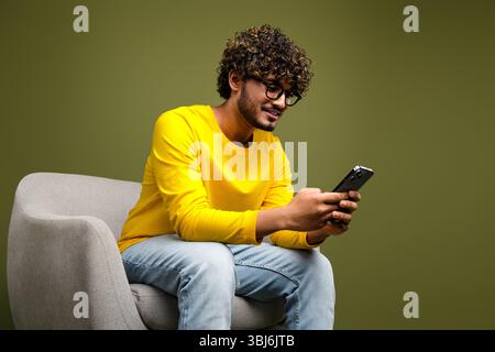 Un jeune homme portant une chemise jaune éclatante utilise un smartphone assis sur une chaise sur un fond neutre Banque D'Images