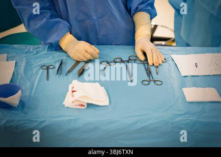 Infirmière assise sur la table d'un instrumentiste dans une salle d'opération de dermatologie, portant des gants et une blouse chirurgicale bleue, en plaçant l'instru chirurgical Banque D'Images