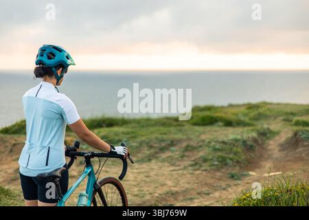 Femme cycliste latine portant casque et gants debout avec son vélo de gravier sur une falaise surplombant la mer au lever du soleil, profitant de la vue paisible et Banque D'Images