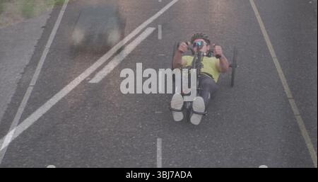 Cycliste couché portant casque en jersey néon et lunettes de soleil sur la route rurale, avec ombre de voiture Banque D'Images