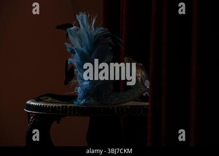 Masque de carnaval repose sur une chaise près d'un miroir et un rideau rouge dans une pièce dans l'obscurité, masque de théâtre Banque D'Images