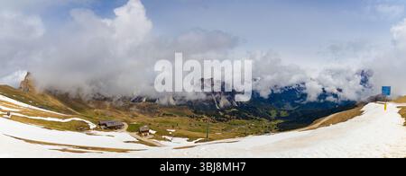 Vue panoramique depuis Seceda, Dolomites, avec des nuages spectaculaires tourbillonnant autour de sommets escarpés, couverture de neige partielle et vallées verdoyantes. Banque D'Images