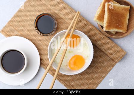 Oeufs à moitié durs dans un bol, sauce soja, baguettes, café et pain grillé sur une table gris clair, pose à plat Banque D'Images