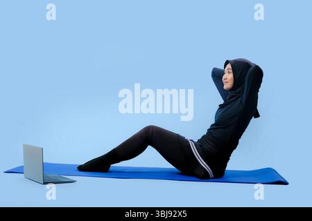 Une femme est assise sur un tapis de yoga avec un ordinateur portable sur ses genoux. Elle porte un top noir et un pantalon noir Banque D'Images