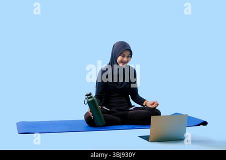 Une femme est assise sur un tapis de yoga avec un ordinateur portable et une bouteille d'eau. Elle est souriante et elle profite de son temps Banque D'Images