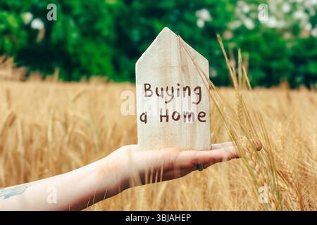 La personne tient un panneau en bois qui dit acheter une maison. Le signe est placé dans un champ de hautes herbes Banque D'Images