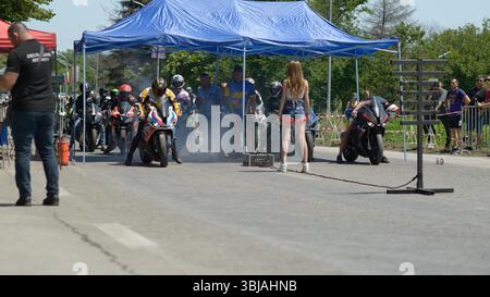 Les motocyclistes se préparent à se lancer sur la ligne de départ d'une course de motos lors du Moto Rock Fest 2025 près de Ruse, en Bulgarie. Plusieurs coureurs en équipement complet et casques sont positionnés sous un auvent bleu, tandis qu'une femme en short se tient devant eux à côté de l'arbre lumineux de départ. De la fumée de pneu est visible lorsqu'un pilote effectue un burn-out. Les spectateurs et l'équipe de l'événement sont vus sur les lignes de touche sous la lumière claire de l'été. L'image capture le moment avant le début de la course sur une section de route délimitée. Banque D'Images