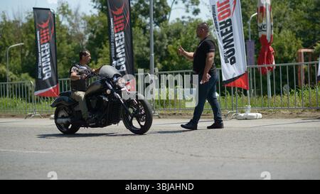 Un motard sur une moto noire mate reçoit un signal pouce levé d'un homme debout dans la rue pendant les préparatifs pour une course de dragsters à l'extérieur de Ruse, Bulgarie. Derrière eux, des drapeaux Power Oil marquent les limites de la zone de traînée. L'image capture le moment avant que le motard ne fasse la queue pour concourir dans le cadre du Moto Rock Fest 2025, qui comprend des activités de course de dragsters. Les rails de sécurité et les conditions estivales sont visibles en arrière-plan. Banque D'Images