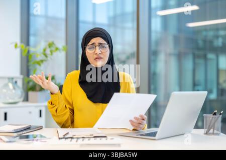 Une musulmane surprise en lunettes regarde des documents dans un bureau, entourée d'outils de travail. Banque D'Images