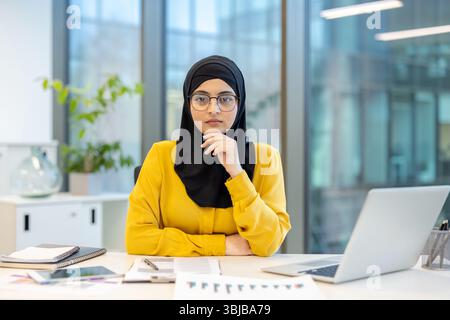 Une femme d'affaires musulmane portant un hijab est assise à son bureau dans un bureau, regardant la caméra avec une expression sérieuse. Banque D'Images